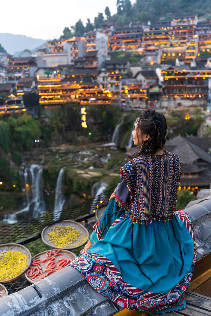 Mujer en traje tradicional chino mirando una ciudad antigua iluminada al anochecer. Ella está sentada en un tejado con vistas a cascadas y edificios tradicionales en una ladera en Lijiang.