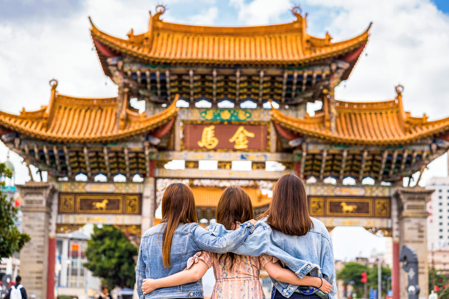 Vista trasera de tres jóvenes abrazadas por la cintura en primer plano. Están frente a la gran y ornamentada Puerta de Oro y Jade (Jinma Bijia) de昆明, China, en una plaza peatonal llena de gente, bajo un cielo nublado.