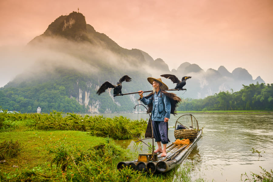 Pescador de cormorán sobre una lancha en el río Yulong en Yangshuo con montañas de fondo. Diseño de viajes a medida en el río Yulong en Yangshuo, China.