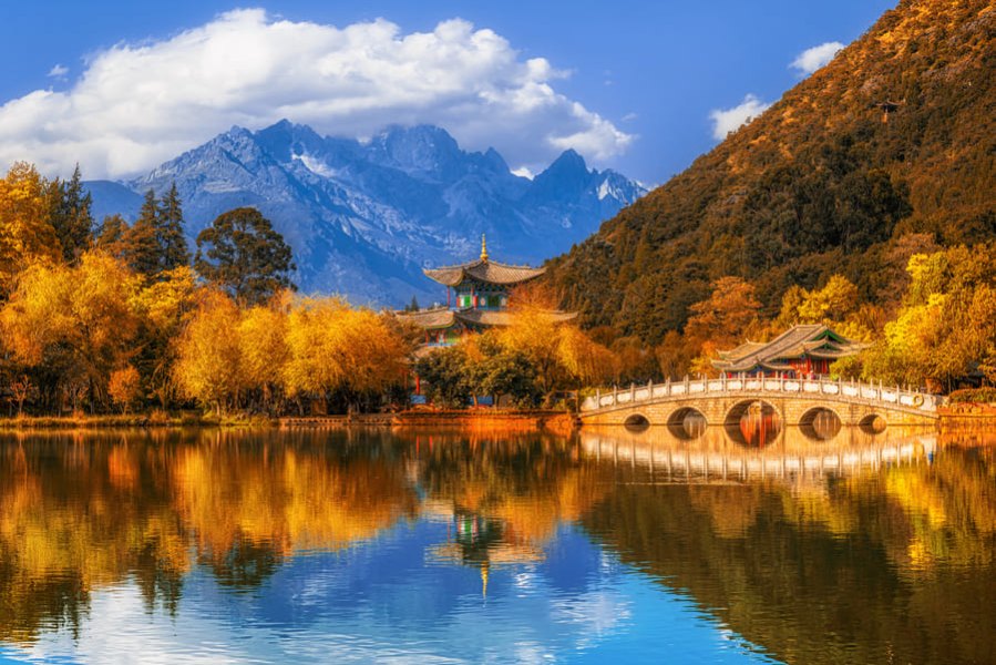 Reflejo de un templo tradicional, un puente arqueado de piedra y árboles dorados de otoño junto a la Montaña de Nieve del Dragón de Jade.