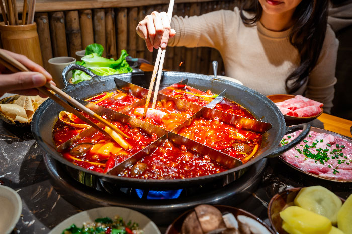 Primer plano de un tradicional hot pot (olla caliente) de Chongqing dividido en varias secciones metálicas sobre una estufa. El caldo es de un color rojo intenso y picante, y se ve a los comensales utilizando palillos para introducir ingredientes frescos como láminas de carne, mientras alrededor de la mesa se disponen platos con verduras, hongos y papas.