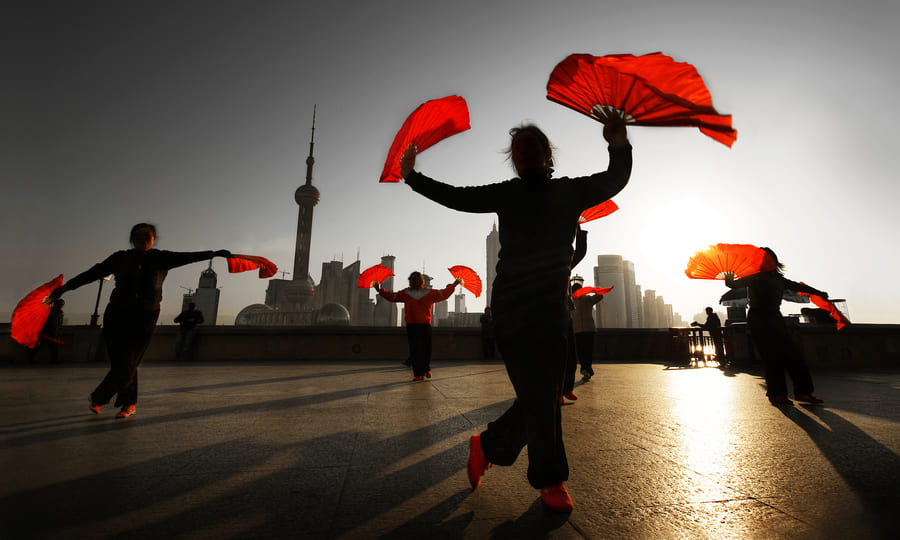 Siluetas de varias personas realizando una danza con abanicos rojos frente al horizonte de Shanghái durante el atardecer. La luz del sol crea un fuerte contraluz y largas sombras sobre el suelo.
