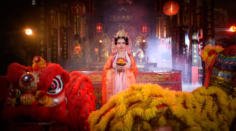 Mujer en traje tradicional sosteniendo mandarinas en el interior de un templo iluminado, flanqueada en primer plano por dos leones de danza tradicional china en color rojo y amarillo.
