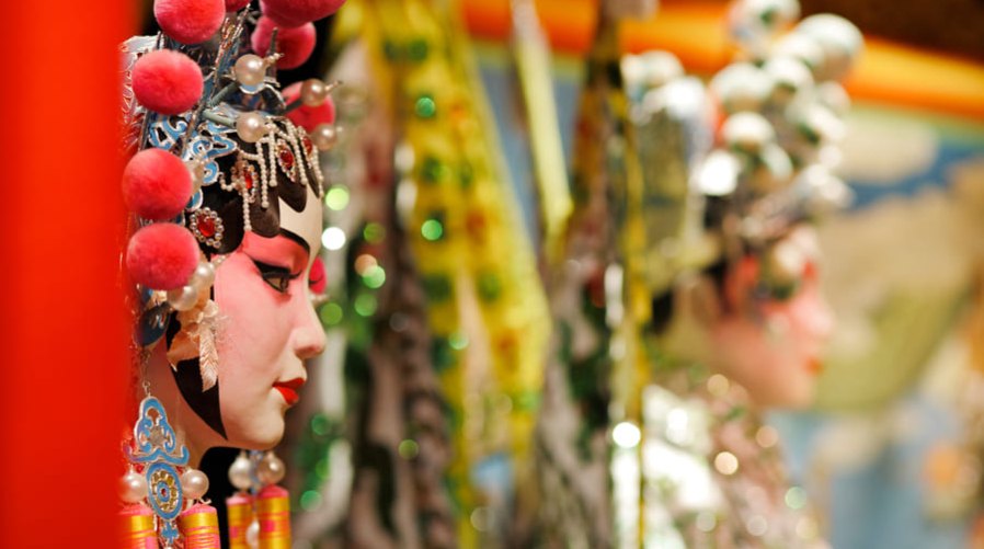 Detalle de perfil de un personaje de la Ópera de Pekín luciendo un tocado festivo con pompones rojos y maquillaje tradicional, con el fondo desenfocado.
