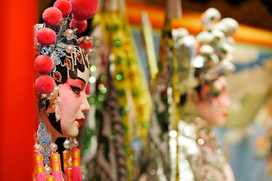 Detalle de perfil de un personaje de la Ópera de Pekín luciendo un tocado festivo con pompones rojos y maquillaje tradicional, con el fondo desenfocado.