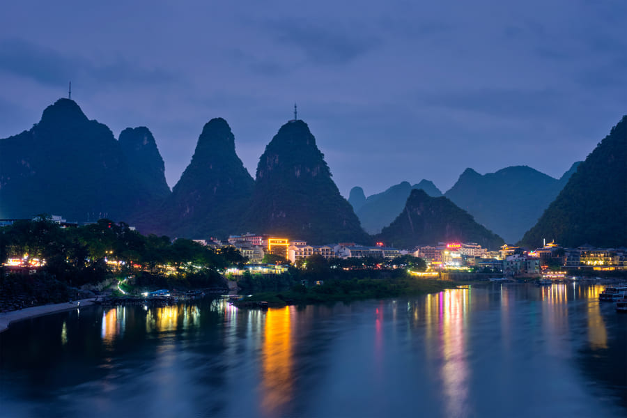 Imagen de una ciudad a orillas de un río en Yangshuo, China, durante la noche. Las luces de la ciudad se reflejan vibrantemente en la superficie del agua. Detrás de los edificios, grandes colinas kársticas cónicas de color verde oscuro se alzan contra un cielo crepuscular de color azul violáceo. Una pequeña isla o banco de arena se ve en el río cerca de la orilla.