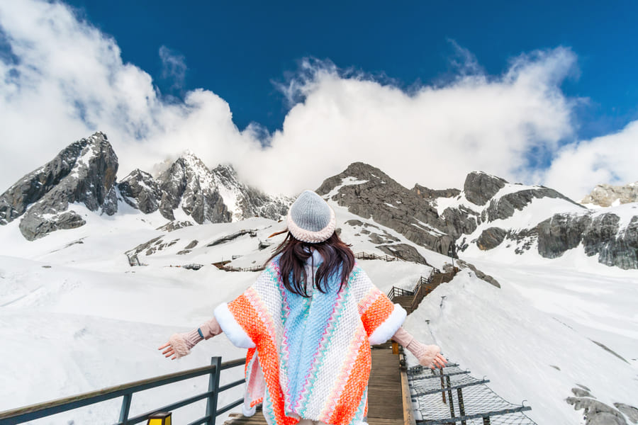 Mujer con poncho multicolor camina por un sendero nevado en la Montaña de Nieve del Dragón de Jade en Yunnan, contemplando los picos alpinos bajo un cielo azul con nubes en Yulongshang, China.