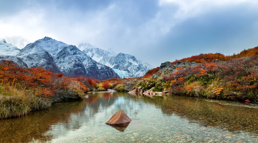 Un río cristalino fluye en primer plano reflejando un paisaje de montaña con picos nevados parcialmente cubiertos por nubes, rodeado de vegetación y arbustos con follaje otoñal en tonos rojizos y naranjas, ideal para ilustrar la naturaleza ancestral en un viaje a Yunnan.