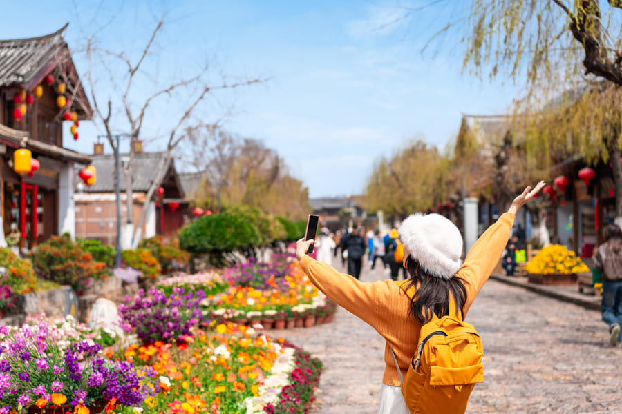 Persona de espaldas fotografiando una calle de arquitectura tradicional en Lijiang. Lleva un sombrero blanco y una mochila amarilla, mientras levanta un brazo frente a un camino decorado con jardines floridos y faroles rojos.